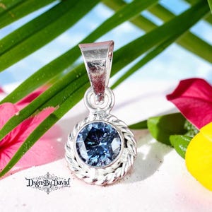 Puede incluir: Un colgante de plata con una piedra preciosa azul redonda y facetada engastada en un borde de cuerda trenzada. El colgante incluye una anilla de plata cónica en la parte superior. El texto DsgrsByDavid está impreso en negro en la esquina inferior.