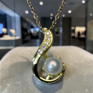 May include: A gold pendant necklace featuring a curved, textured gold frame accented with a row of small, clear, sparkling stones. A single, round, white pearl is nestled within the lower curve of the gold setting, suspended from a delicate gold chain.