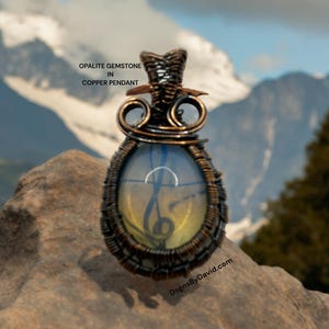 Könnte beinhalten: Opal-Edelstein in einem kupferdrahtgewickelten Anhänger. Der Anhänger hat eine Tropfenform und der Edelstein ist hellblau und gelb. Der Anhänger liegt auf einem Stein mit einer Bergkette im Hintergrund.