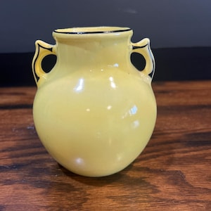 Könnte beinhalten: Eine kleine, gelbe Keramikvase mit schwarzem Besatz und zwei Henkeln. Die Vase ist etwa 10 cm hoch.