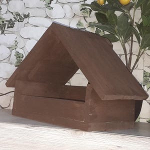 Könnte beinhalten: Ein braunes Vogelhaus aus Holz mit einem Satteldach. Das Vogelhaus hat eine rechteckige Basis und ein einfaches, offenes Design. Es steht auf einem Regal, mit einem dekorativen Hintergrund aus einer Pflanze und einer Steinmauer.