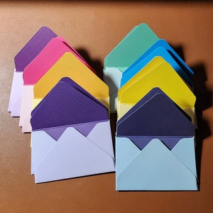 May include: A stack of colorful envelopes in various shades of blue, purple, yellow, pink, and green. The envelopes are arranged in a fan shape, with the top envelope being a light blue color.