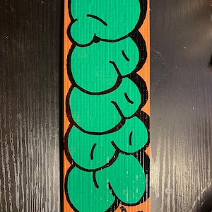 May include: A green and black graffiti art piece on a rectangular orange background. The art is made up of several bubble letters, each with a black outline and a green interior. The letters are stacked on top of each other, creating a vertical design. The piece is signed with the text "#678 #33 #33".