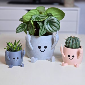 Könnte beinhalten: Drei graue und rosafarbene Keramik-Blumentöpfe mit Comic-Gesichtern und Armen. Die Töpfe halten kleine Pflanzen. Die grauen Töpfe haben schwarze Augen und einen lächelnden Mund. Der rosafarbene Topf hat schwarze Augen und einen lächelnden Mund.