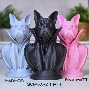 Könnte beinhalten: Drei 3D-gedruckte Büsten eines französischen Bulldoggen, die einen Dracula-Umhang tragen. Die Büsten sind in drei verschiedenen Farben: hellblau, schwarz und rosa. Der Text unter jeder Büste lautet "Marmor", "Schwarz Matt" und "Pink Matt".