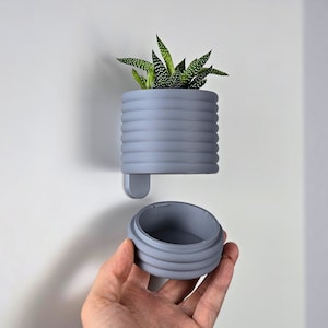 Könnte beinhalten: Ein grauer Pflanzer mit einer Sukkulentenpflanze darin. Der Pflanzer besteht aus einem gerippten Material und hat einen abnehmbaren Deckel.