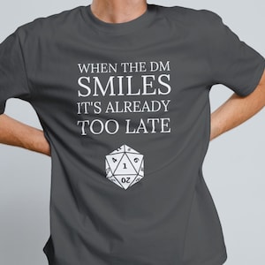 Puede incluir: Una camiseta gris oscuro con el texto "When the DM smiles it's already too late" y un gráfico de dado de 20 caras blanco.