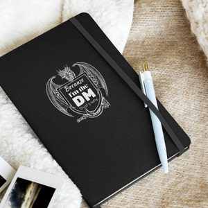Puede incluir: Cuaderno negro con un diseño de dragón blanco y el texto "Because I'm the DM. That's why."