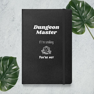 May include: Black notebook with a white inscription that reads "Dungeon Master" and "If I'm smiling, you're not". A white graphic of a dice with a skull and crossbones is in the center of the notebook.