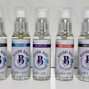May include: A row of nine clear glass spray bottles with silver pump tops. Each bottle features a white label with the text Pleasure Blast, a large letter B, and a unique scent name such as Exotic Island Girl, Seduced, Secret Pleasure, and Black Ice.