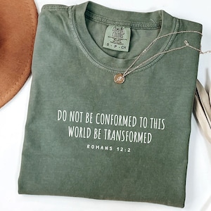 Peut inclure: Un t-shirt vert avec le texte "DO NOT BE CONFORMED TO THIS WORLD BE TRANSFORMED" et "ROMANS 12:2" en blanc. Le t-shirt est plié et posé à plat sur une surface blanche.