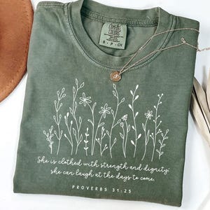 May include: Moss green t-shirt with white floral design and the text "She is clothed with strength and dignity; she can laugh at the days to come. PROVERBS 31:25". A gold necklace with a pendant is on the shirt.