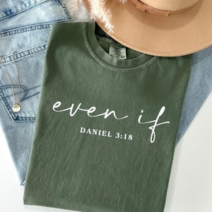 May include: A green t-shirt with the text "even if" and "DANIEL 3:18" in white lettering. The shirt is folded and laid on a light blue denim background.