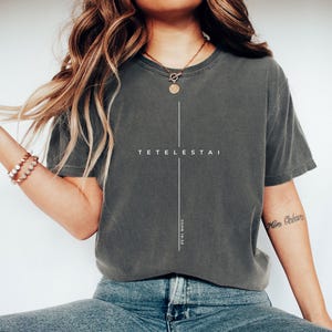 May include: Dark gray t-shirt with the word "TETELESTAI" and a vertical line design. The text "JOHN 19:30" is printed vertically on the lower right side of the shirt. The model is wearing blue jeans and white shoes.