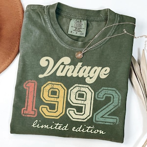 May include: Olive green t-shirt with the text "Vintage 1992 limited edition" in a retro font. The numbers are in a mix of colors: red, yellow, and blue. A gold necklace with a pendant is on the shirt.