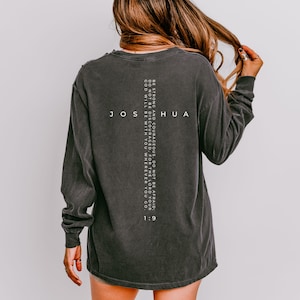 May include: Dark gray long-sleeve shirt with the word "JOSHUA" and the Bible verse Joshua 1:9 printed in white. The shirt has a distressed look and a relaxed fit.