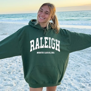May include: A dark green hoodie featuring the white text "RALEIGH" above "NORTH CAROLINA". The hoodie has a front pocket and a drawstring hood. The person is wearing the hoodie on a sandy beach with the sea in the background.