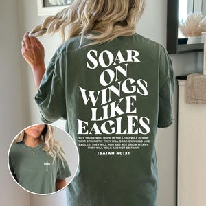 May include: Olive green t-shirt with the text "SOAR ON WINGS LIKE EAGLES" in white on the back. The shirt also features a small white cross on the front. The text "ISAIAH 40:31" is also printed on the back.