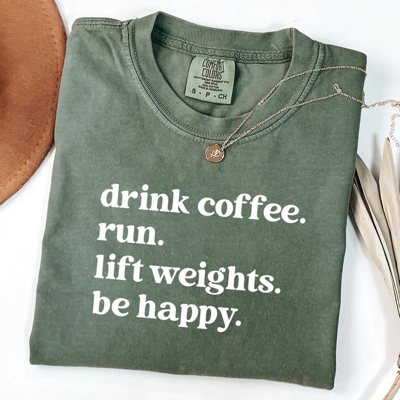 May include: An olive green Comfort Colors t-shirt with the text "drink coffee. run. lift weights. be happy." in white. A gold-colored necklace with a pendant is on the shirt.