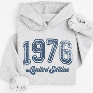 May include: A light gray hoodie with the year "1976" in distressed navy blue lettering and the words "Limited Edition" below. The hoodie has a drawstring hood and ribbed cuffs.
