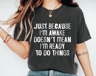 May include: Dark gray t-shirt with the text "JUST BECAUSE I'M AWAKE DOESN'T MEAN I'M READY TO DO THINGS" in white. The shirt is worn with blue jeans and a gold necklace.