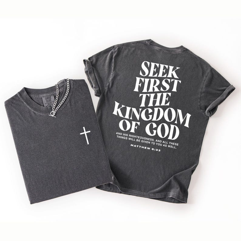 May include: Two charcoal grey t-shirts. One is folded with a white cross and a silver chain necklace. The other shirt has the text "SEEK FIRST THE KINGDOM OF GOD" and a Bible verse printed on the back. The shirts are on a white background.