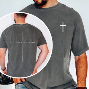 May include: A charcoal gray short-sleeve t-shirt featuring a small white cross on the front left chest. The back of the shirt displays a large vertical white cross with the text "FATHERHOOD IS KINGDOM WORK" printed horizontally across the center.