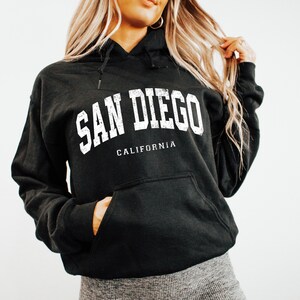May include: Black hooded sweatshirt with the text "SAN DIEGO CALIFORNIA" in white block letters. The sweatshirt has a front pocket and a drawstring hood. The garment is made of a soft, comfortable material.