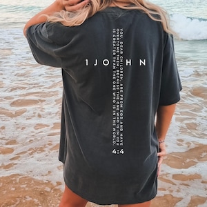 Puede incluir: Camiseta gris oscuro con texto blanco en la espalda. El texto incluye "1 JOHN" y un versículo bíblico: "USTEDES, HIJOS, SON DE DIOS Y LOS HAN VENCIDO, PORQUE EL QUE ESTÁ EN USTEDES ES MAYOR QUE EL QUE ESTÁ EN EL MUNDO. 4:4."