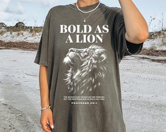 May include: Olive green t-shirt with the text "BOLD AS A LION" above a detailed lion illustration. The shirt also features a Bible verse, Proverbs 28:1, in white text. The shirt is a relaxed fit.