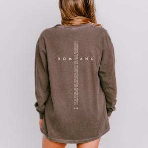 May include: A long-sleeve brown t-shirt featuring white text on the back. The word ROMANS is printed horizontally, intersected by a vertical cross containing the scripture: I CONSIDER THAT OUR PRESENT SUFFERINGS ARE NOT WORTH COMPARING WITH THE GLORY THAT WILL BE REVEALED IN US. 8:18.