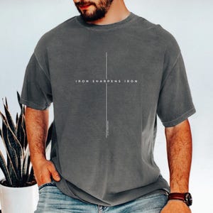 May include: A gray t-shirt with the text "IRON SHARPENS IRON" in white, centered on the chest. A vertical white line runs from the text to the bottom of the shirt. The text "PROVERBS 27:17" is printed vertically below the line.