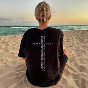 May include: A person wearing a black t-shirt with the text "TRUST IN THE LORD WITH ALL YOUR HEART, AND LEAN NOT ON YOUR OWN UNDERSTANDING, IN ALL YOUR WAYS ACKNOWLEDGE HIM, AND HE SHALL DIRECT YOUR PATHS 3:5-6" printed on the back. The person is sitting on a sandy beach with the sea in the background.