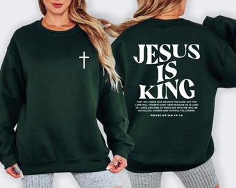 May include: A dark green crewneck sweatshirt featuring a small white cross on the left chest. The back displays the white text "JESUS IS KING" in large letters, followed by the verse "THEY WILL WAGE WAR AGAINST THE LAMB" and "REVELATION 17:14".