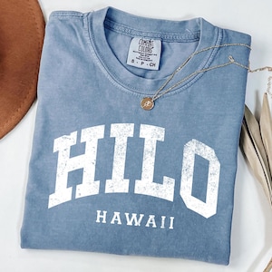 May include: A blue Comfort Colors t-shirt with the word "HILO" in white block letters and "HAWAII" underneath. A gold necklace with a pendant rests on the shirt. A brown hat is partially visible in the upper left corner.
