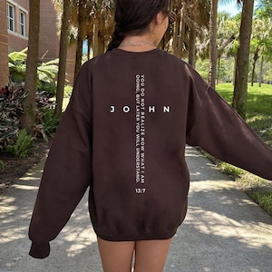 May include: A dark brown sweatshirt with the word "JOHN" vertically aligned and the text "YOU DO NOT REALIZE NOW WHAT I AM DOING, BUT LATER YOU WILL UNDERSTAND. 13:7" printed in white. The sweatshirt is a long-sleeved, casual garment.