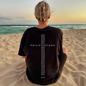 May include: A person wearing a black t-shirt with the text "PHILIPPIANS" and a Bible verse printed on the back. The person is sitting on a sandy beach, facing the ocean. A light-colored hair clip is in their hair.