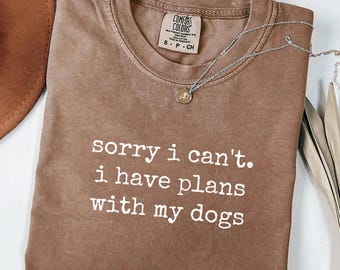 May include: A brown Comfort Colors t-shirt with the text "sorry i can't. i have plans with my dogs" in white. A silver necklace with a small pendant is draped across the shirt.