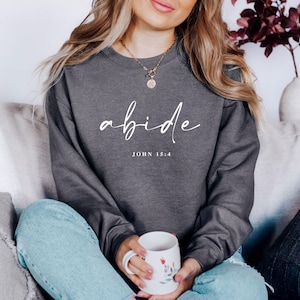 May include: A charcoal gray sweatshirt with the word "abide" in white script, and the text "JOHN 15:4" below it. The sweatshirt is worn with light blue jeans. A white mug with floral designs is held in the hands.