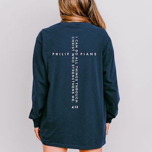 May include: Navy blue long-sleeve shirt with white text in a cross design. The text reads "PHILIPPIANS" and "I CAN DO ALL THINGS THROUGH CHRIST WHO STRENGTHENS ME. 4:13". The shirt is a casual style.
