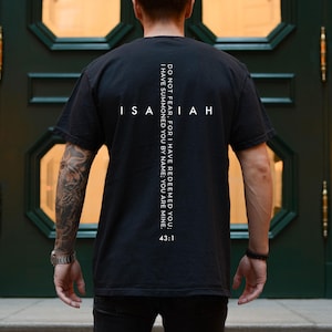 May include: Black t-shirt with the word "ISAIAH" in white, with a vertical line of text that reads "DO NOT FEAR, FOR I HAVE REDEEMED YOU; I HAVE SUMMONED YOU BY NAME; YOU ARE MINE. 43:1". The shirt is worn by a person.
