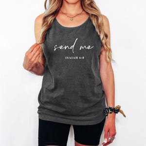 May include: Dark gray tank top with the words "send me" in a cursive font, and the text "ISAIAH 6:8" below. The top is worn by a person with long blonde hair, black shorts, and jewelry.
