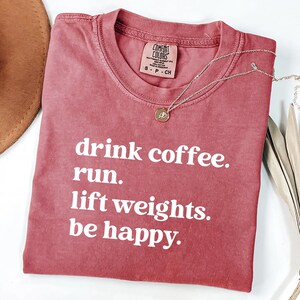 May include: A dusty rose Comfort Colors t-shirt with the phrases "drink coffee. run. lift weights. be happy." printed in white. A silver necklace with a circular pendant rests on the shirt.