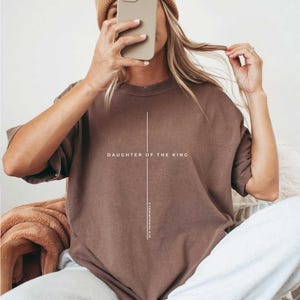 May include: A brown short-sleeved t-shirt with the text "DAUGHTER OF THE KING" and "2 CORINTHIANS 6:18" printed in white. The shirt has a vertical line design. The shirt is a neutral color.
