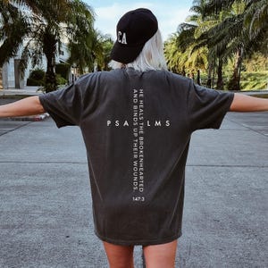 May include: A charcoal gray short-sleeve t-shirt featuring white text on the back arranged in a cross shape. The text reads PSALMS, HE HEALS THE BROKENHEARTED AND BINDS UP THEIR WOUNDS, and 147:3. A black baseball cap with the letters CA is also visible.