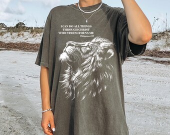 May include: Dark olive green t-shirt featuring a white lion graphic and the text "I CAN DO ALL THINGS THROUGH CHRIST WHO STRENGTHENS ME" from Philippians 4:13. The shirt has a relaxed fit and short sleeves.