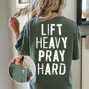 May include: Olive green t-shirt with the words "LIFT HEAVY PRAY HARD" printed in white. A small circular image shows a person wearing a similar shirt with a cross pendant.