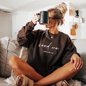 May include: A woman wearing a brown sweatshirt with the text "send me" and "Isaiah 6:8" in white script. She is holding a black mug and wearing fuzzy slippers while sitting on a gray couch.