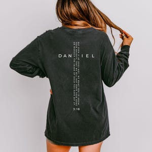 May include: Dark grey long-sleeved shirt with the word "DANIEL" and the text "BUT EVEN IF HE DOES NOT, WE WANT YOU TO KNOW. YOUR MAJESTY, THAT WE WILL NOT SERVE YOUR GODS OF WORSHIP THE IMAGE OF GOLD YOU HAVE SET UP. 3:18" printed on the back.
