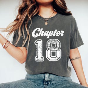 Peut inclure: T-shirt gris foncé avec l'inscription blanche "Chapter 18" en lettres usées. Le chiffre 18 est dans un style de lettres en bloc. Le t-shirt est de style décontracté et unisexe.